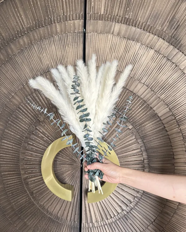 A hand holding a bouquet of white pampas grass and eucalyptus in front of a carved wood panel at Meridian Acupuncture