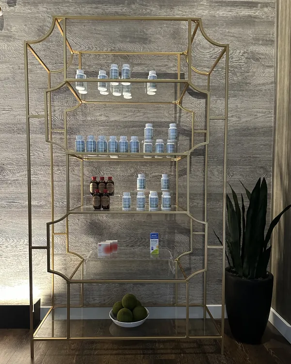 A gold and glass display shelf stocked with bottles of herbal formulas at Meridian Acupuncture, beside a potted plant